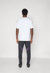 Witte kortebloezende t-shirt met zwarte schouderaccenten; gecombineerd met zwarte broek met paarse diagonale strepen en witte zijstrepen.
