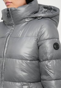Harmaa puffatakki, jossa on kiiltävä pinta, korkea kaulus ja nyörihuppu. Vasemmassa hihassa on musta logo-patch.
