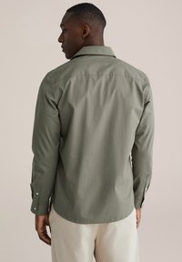 Olivno zelena majica z dolgimi rokavi, ki ima klasičen ovratnik, gumbe na manšetah ter gladko teksturo, rahlo prilegajoč kroj in ukrivljen rob.