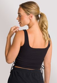 Zwarte geribbelde cropped top met brede bandjes en een vierkante achterkant; heeft zij-koordjes voor een verstelbare pasvorm. Gekleed met donkere broek.