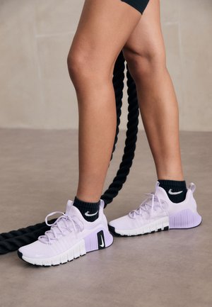 Light purple athletic shoes with a textured upper, white and black sole, and logo accents. Worn with black knit socks and a workout backdrop.