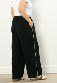 Svarta vida byxor med elastisk midja, med ett vitt sidospel och sidofickor. Matchas med vita slip-on skor.
