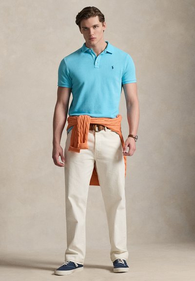 Νεαρός άνδρας όρθιος, φοράει ανοιχτό γαλάζιο πόλο, μπεζ παντελόνι, μπλε σκούρα αθλητικά παπούτσια, πορτοκαλί πουλόβερ δεμένο γύρω από τη μέση, ουδέτερο φόντο.