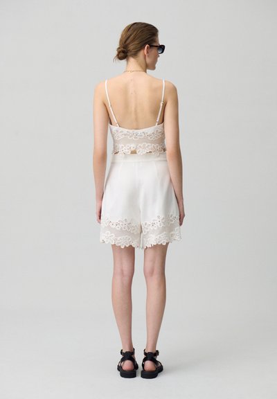 Completo a due pezzi composto da un top corto in pizzo bianco con spalline sottili e pantaloni shorts abbinati con orlo in pizzo. Indossato con sandali neri.