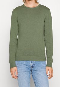 Olijfgroen gebreide trui met een ronde hals, lange mouwen en geribde manchetten. Gecombineerd met lichtblauwe denim jeans.