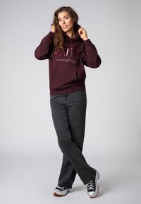 Bordeaux hoodie met tekst, gemusterde trekkoorden en een voorzak, gecombineerd met donkergrijze jeans en zwart-witte sneakers.