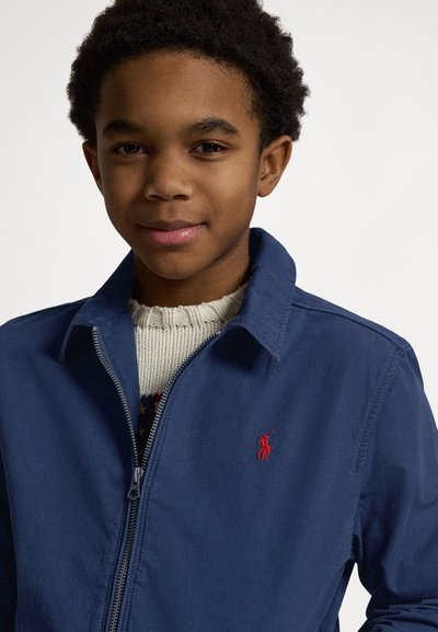 Tamsiai mėlynas užsegamas švarkelis iš lengvos medžiagos. Kairėje krūtinės pusėje yra raudonas logotipo emblema ir apykaklė. Po juo dėvimas tekstūruotas megztinis.