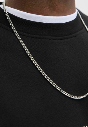 Felpa nera con un collo alto, sovrapposta a un colletto bianco. Una collana di catena argentata con maglie intrecciate si appoggia sul tessuto.