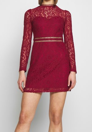 Femme portant une mini-robe en dentelle bordeaux ajustée, à manches longues, col montant, avec un détail de taille découpé en cercle.