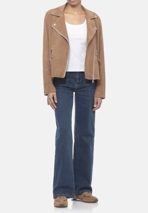 Donna con giacca biker in camoscio marrone, top bianco, jeans svasati blu e sneakers marroni, in piedi su sfondo bianco.