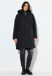 Femme portant un manteau d'hiver noir à capuche, un col roulé gris, un jean noir délavé et des bottines noires, se tenant contre un fond blanc.