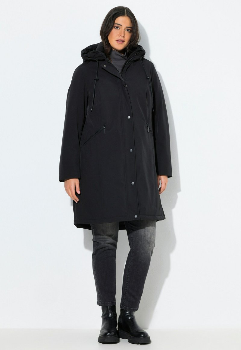 Femme portant un manteau d'hiver noir à capuche, un col roulé gris, un jean noir délavé et des bottines noires, se tenant contre un fond blanc.
