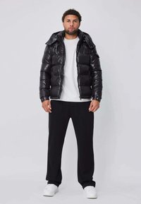 Veste matelassée noire à finition brillante, dotée d'un col haut et d'une fermeture éclair, portée avec un t-shirt blanc et un pantalon noir.