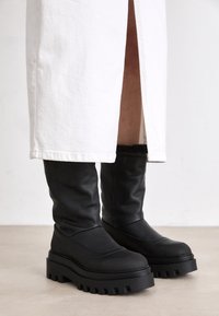 Botas de cuero negro hasta la rodilla con una suela de goma gruesa y paneles texturizados, con un acabado suave y estrías elevadas a lo largo de la suela.