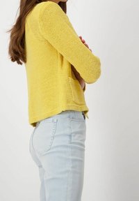 Gul stickad cardigan med långa ärmar och en framficka, ihopparad med ljusblå denimjeans, som visar en texturerad tyg.