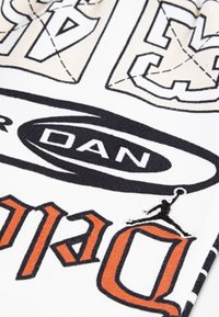 Tissu blanc présentant des imprimés graphiques noirs et oranges, y compris un logo avec la silhouette d'un joueur de basket en plein saut.
