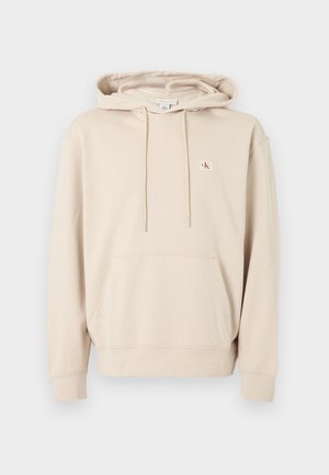 Béžová mikina s kapucí, přední kapsa na vak, šňůrky, žebrované manžety a malé červenobílé logo CK na levé straně hrudi.