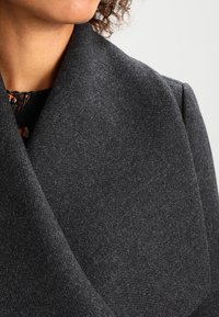 Cappotto di lana grigio scuro con una texture morbida e un ampio colletto, caratterizzato da un design pulito e su misura. Il tessuto appare caldo e strutturato.