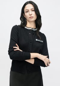 Črni kardigan z okroglim izrezom, okrašen s prosojnim ovratnikom in poudarki na žepih z bleščicami. Dizajn izstopa s pleteno teksturo in gumbi za zapenjanje.