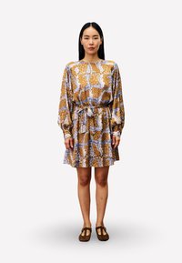 Bedrucktes Kleid mit langen Ärmeln, braun, blau und weißem Blumenmuster, tailliert, mit ausgestelltem Rock, kombiniert mit braunen flachen Schuhen.