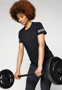 Camisola atlética preta de mangas curtas com a marca "BORG" nas mangas, combinada com leggings escuras, enquanto uma pessoa realiza um exercício de levantamento de peso.
