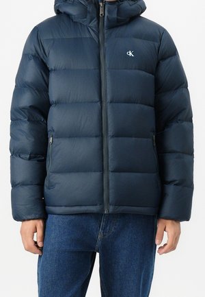 Persoon draagt een donkerblauwe doorgestikte pufferjas met ritssluiting aan de voorkant, zijzakken met rits en een klein "cK"-logo op de borst, gecombineerd met een blauwe spijkerbroek.