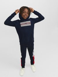 Mornarska dukserica s kapuljačom s prednjim grafičkim uzorkom koji uključuje pruge i natpis "JACK & JONES", u kombinaciji s mornarskim trenerkama i bijelim tenisicama.