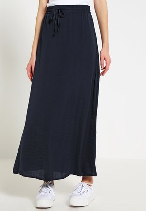 Femme portant une longue jupe noire à taille coulissée et des baskets blanches, debout devant un fond clair uni.
