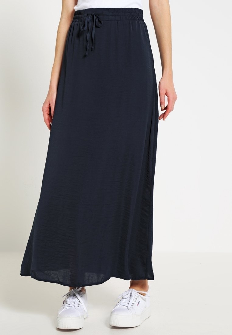 Femme portant une longue jupe noire à taille coulissée et des baskets blanches, debout devant un fond clair uni.
