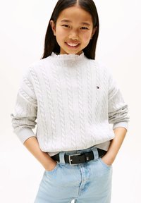 Hellgrauer Strickpullover mit Zopfmuster und hohem, geripptem Kragen, kombiniert mit blauen Jeans und einem schwarzen Gürtel mit silberner Schnalle.