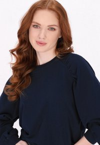 Felpa blu navy con maniche arricciate, tessuto morbido, scollo a girocollo e vestibilità rilassata, indossata da una persona con lunghi capelli ondulati di colore rosso-marrone.