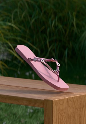 Pink klipklap med blomsterdekorationer balancerende på kanten af et træbord udendørs med grønne planter sløret i baggrunden.