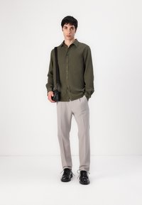 Camisa verde oliva tipo botonera, pantalones beige claros, zapatos negros y un pequeño bolso cruzado negro. Diseño simple con un ajuste relajado.