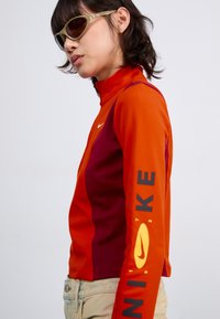 Langermet Nike-genser med livlige oransje- og dype burgunderpaneler, glidelås i kragen og dristig logodetalj i gult langs ermet.