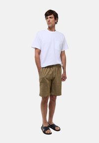 T-shirt blanche à manches courtes avec une poche, associée à un short brun à motifs et des sandales noires. Look décontracté, éléments de design minimalistes.