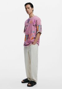 Roze oversized t-shirt met abstracte kleurrijke prints, gecombineerd met crèmewitte gestreepte wijde broek en zwarte instapsandalen.