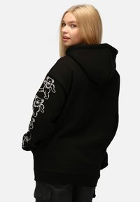 Felpa nera con cappuccio e tasca a marsupio, caratterizzata da illustrazioni di orsi bianchi sulla manica sinistra. Tessuto spesso con finitura testurizzata e cappuccio.