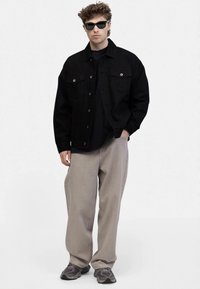 Chaqueta de mezclilla negra con bolsillos abotonados, superpuesta sobre una camisa negra. Pantalones sueltos gris claro y zapatillas oscuras completan el look. Se llevan gafas de sol.