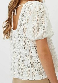 Top bianco in broderie anglaise con maniche a palloncino e schiena bassa, caratterizzato da intricate fantasie a motivo intagliato e una finitura morbida e testurizzata.
