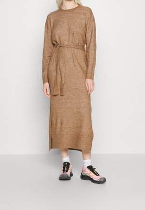 Kobieta w długiej brązowej dzianinowej sukience z paskiem do wiązania, białe skarpetki i różowo-czarne buty sportowe, stojąca na tle białej ściany.