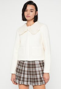 Λευκό σακάκι από μείγμα μαλλιού με υφή και μεγάλο πλεκτό γιακά, δύο μπροστινές τσέπες, συνοδευόμενο από πιετέ φούστα καρό σε καφέ και λευκό.