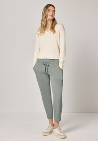 Kremowy sweter z dekoltem w serek, szare spodnie dresowe z troczkiem, z dwoma przednimi kieszeniami oraz jasne sneakersy. Miękki materiał o luźnym kroju i zwężanych nogawkach.