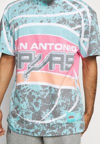 Mand iført en farverig San Antonio Spurs T-shirt med et abstrakt basketballdesign i blå, pink, orange og grå.