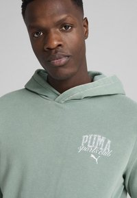 Sudadera verde clara hecha de un tejido suave, con un bolsillo tipo canguro, capucha con cordón y un logo impreso en blanco en el lado izquierdo del pecho.