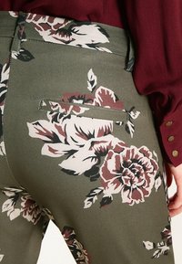 Kalhoty s květinovým vzorem v olivově zelené látce, s velkými růžovými a burgundskými květinami. Obsahují zadní kapsu a plochý design pasu.