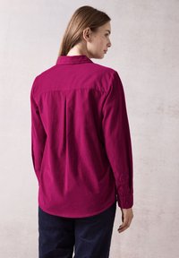 Magenta long-sleeve shirt with a collared neckline and back pleat detail, made of a smooth fabric. Paired with dark, loose-fitting trousers.