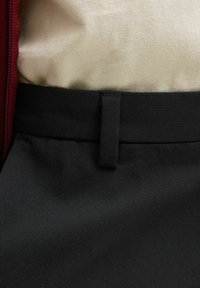 Chemise beige rentrée dans un pantalon noir avec passants pour ceinture, veste rouge à fermeture éclair partiellement visible sur le côté gauche.