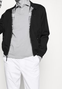 Černá bomber bunda s károvanou podšívkou, šedý polo tričko a bílé kalhoty. Obsahuje zip vpředu a boční kapsy. Hladká textura a strukturovaný střih.