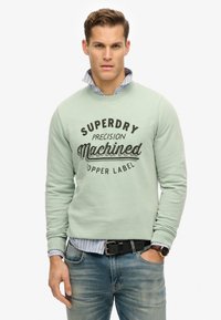 Sweat-shirt vert clair avec le texte imprim é en noir "SUPERDRY Precision Machined." Porté par-dessus une chemise à rayures bleues et blanches ; jeans en denim visibles.