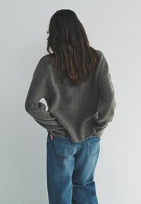 Sivý pletený sveter s voľným strihom a rebrovanými manžetami, skombinovaný s svetlomodrými voľnými džínsami. Textúrovaná tkanina pridáva hĺbku.